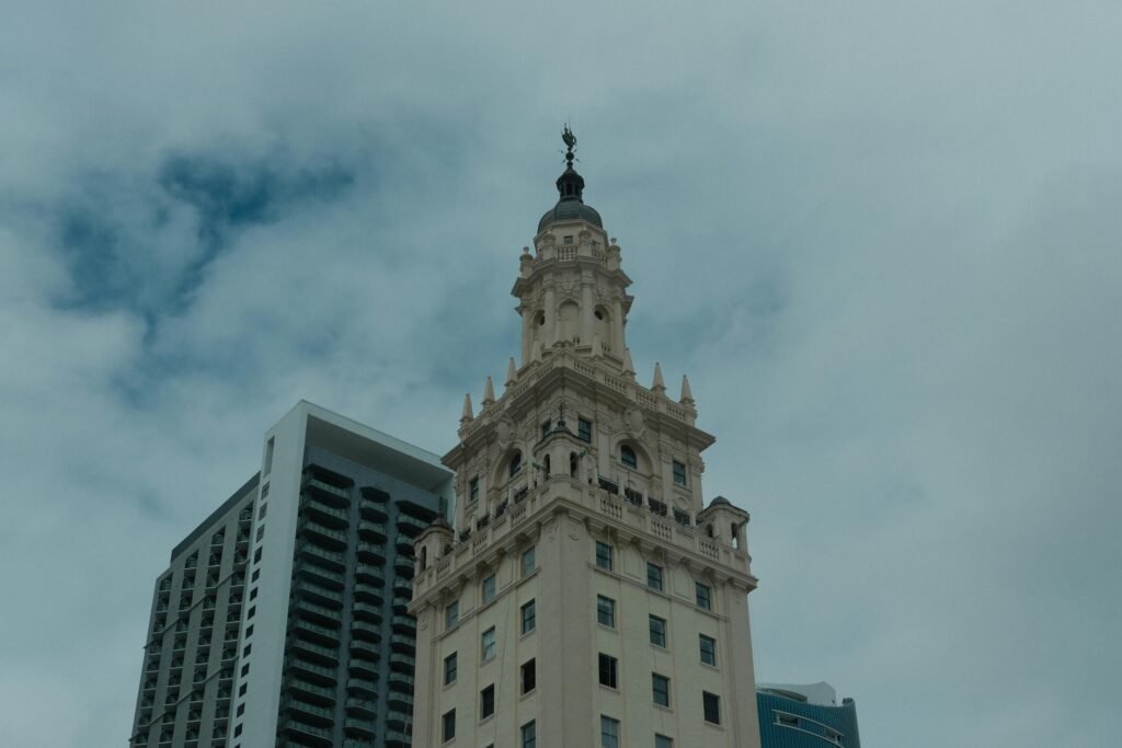 A historic tower stands prominently against a cloudy sky with modern buildings in the background.