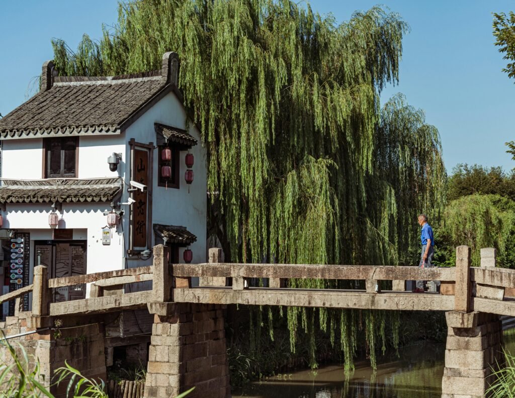 A serene scene featuring traditional Chinese architecture and a man crossing a stone bridge under willow trees.